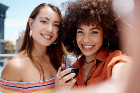 Two beautiful mix race women smiling taking a selfie while enjoying a glass of red wine while...