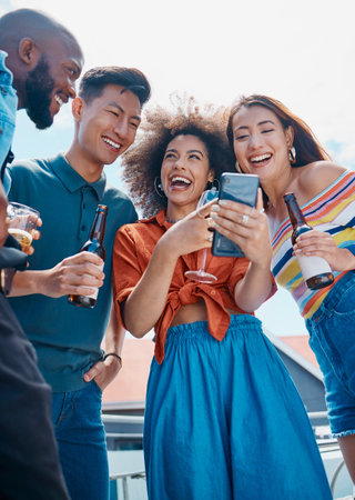 A group of diverse young cheerful friends standing outside together and using a cellphone for selfies and social media. Smiling men and women being social and celebrating on a weekend partyの写真素材