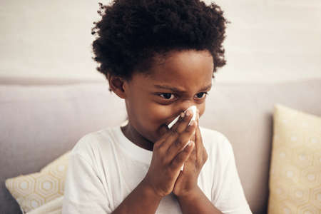 Cold and flu season. Sick african american boy with afro blowing nose into tissue. Child suffering from a runny nose or sneezing, covering his nose while sitting at home. Time to get vaccinatedの写真素材