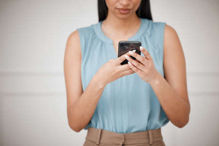 Closeup of a young formal businesswoman using a cellphone to text standing against a white background. Mixed race female checking social media .Corporate worker browsing the internet for researchの写真素材