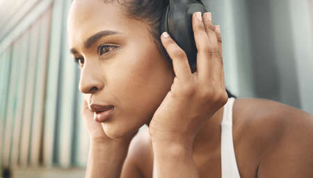Closeup of one fit young hispanic woman listening to music with headphones while exercising in an urban setting outdoors. Face of focused and motivated female athlete ready for training workout or runの写真素材