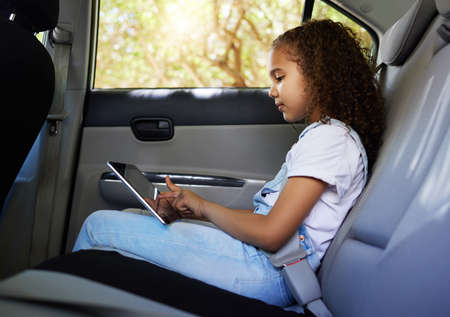 Her travel tablet. Cropped shot of an adorable little girl using her tablet while sitting in the backseat of a car.の写真素材