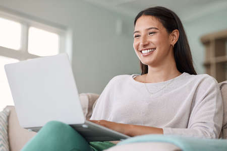 Shot of a beautiful young woman using her laptop while sitting on the couch at home.の写真素材