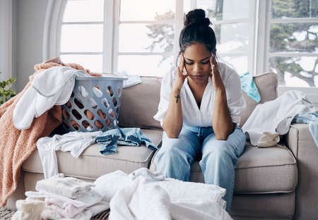 Mothers dont get a break. Shot of an attractive young woman sitting alone in her living room and feeling stressed wile doing the laundry.の写真素材