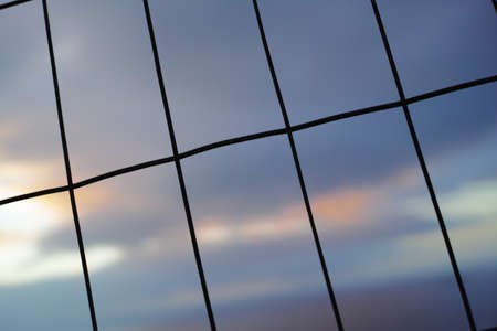 Iron fence in vertical, horizontal lines against a blur sky background. Wire meshed in rectangle shapes for security or safety concept. The texture of window patterns or dark silhouettes of a gateの写真素材
