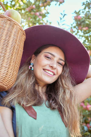 Portrait of one happy woman holding basket of fresh picked apples from trees on sustainable orchard farm outside on sunny day. Face of cheerful farmer harvesting juicy organic fruit in season to eatの写真素材