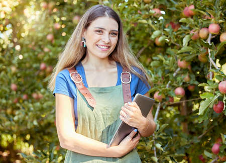 Portrait of a female farmer holding a digital tablet while standing by apple trees. Smiling woman...