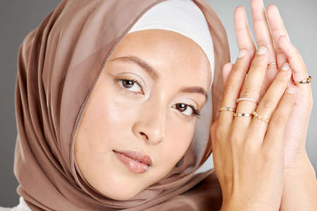 Studio portrait of beautiful muslim woman wearing a brown headscarf and showing multiple rings on her hands. Female wearing hijab and glowing makeup with trendy jewelry showing off her modest beautyの写真素材