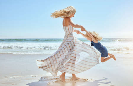 Happy mother swinging and spinning cute daughter in circles by the arms at the beach. Playful, energetic and joyful kid having fun while bonding with mom on sunny summer vacation outdoorsの写真素材