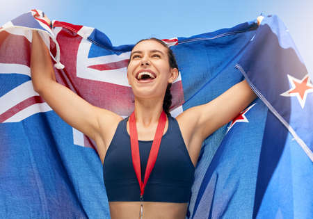 Young fit female athlete cheering and holding a New Zealand flag with a gold medal after competing in the Olympics. Smiling active sporty woman feeling motivated and celebrating winning in sportの写真素材