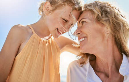 Cheerful mature woman and little girl talking and sharing a secret while sitting on the beach. Happy little girl smiling while sitting with her mom or grandmother and being loving and affectionateの写真素材