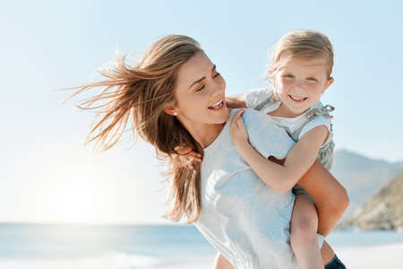 I just want her ti have the best childhood. Shot of a woman carrying her daughter on her back while at the beach.の写真素材