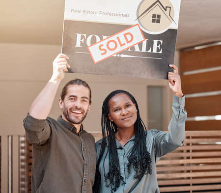 We did it. Shot of a young couple holding up a sold board outside their house.の写真素材