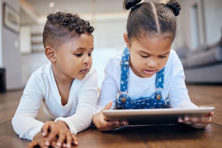 Two young mixed race children using a digital tablet while lying together on the floor at home. Young boy and girl siblings browsing the internet online to play games and learn from educational appsの写真素材