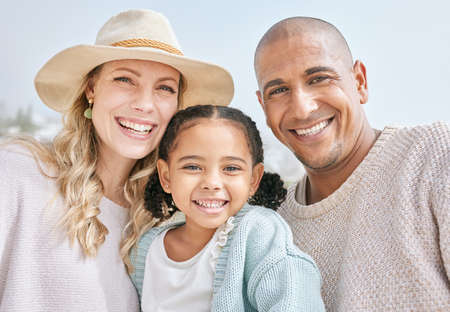 Portrait of an interracial happy family bonding while on a beach vacation together. Cute little girl embracing and sitting with her parents outside, enjoying fresh summer air with copyspaceの写真素材