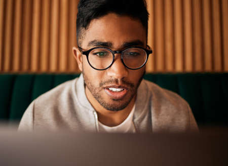 .. Closeup of geeky young indian man wearing glasses while reading something interesting and sitting inside. Man wearing glasses while reading online. Dedicated male student doing research in cafe.の写真素材