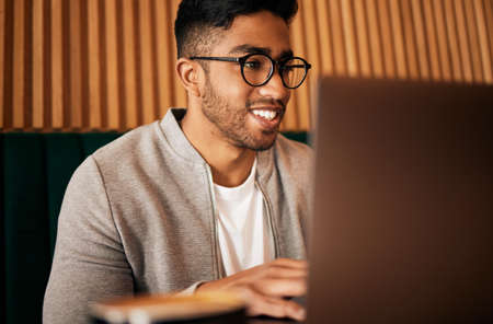 .. Handsome indian man with glasses sitting alone and using laptop in cafe. Man using wifi to browse...