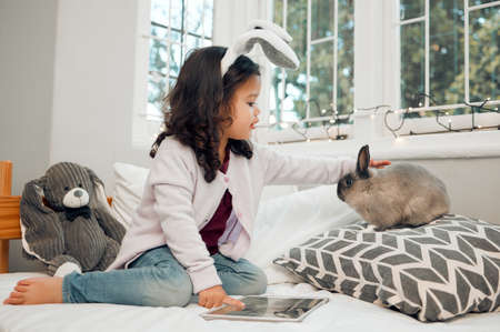 My fluffy friend. Shot of an adorable little girl using a digital tablet and bonding with her pet rabbit on her bed.の写真素材