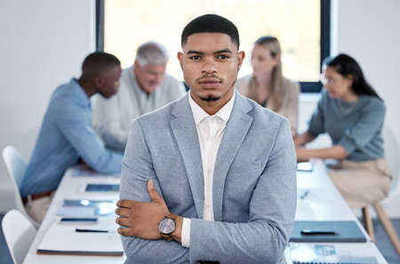I am not afraid to take risks where needed. Portrait of a confident young businessman standing with his arms crossed in an office while his colleagues have a meeting in the background.の写真素材
