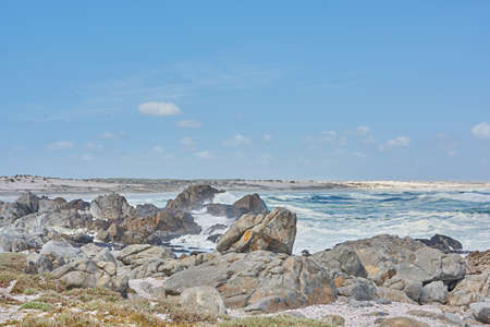 A shallow rocky coastline in the Cape Province, South Africa. Ocean waves crashing on coastal rocks and boulders on a sunny summer day, blue clear skies, and a scenic tropical landscape beachfrontの写真素材
