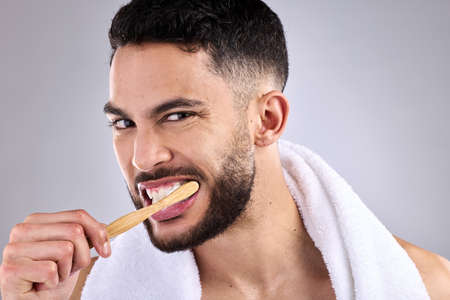 Be gentle but thorough. a young man brushing his teeth while standing against a grey background.