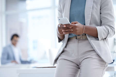 Taking note of all updates that are happening. Closeup shot of an unrecognisable businesswoman using a cellphone in an office with her colleague in the background.の写真素材