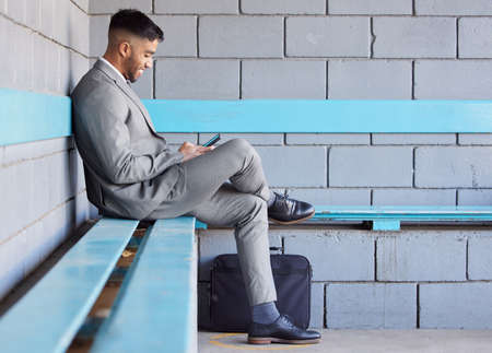 Staying mobile during his morning commute. a young businessman using a cellphone while sitting on a bench at a railway station during his commute.の写真素材