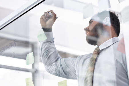 He has the plan all laid out. Low angle shot of a young businessman writing notes on a glass wall in...