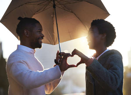 Their love blooms throughout all seasons. a young man and woman making a heart shape with their hands while standing under an umbrella together on a rainy day in the city.の写真素材