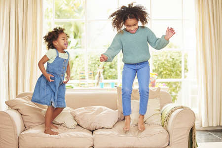 Who can jump the highest. Full length shot of two adorable girls jumping on the sofa in the living room at home.の写真素材