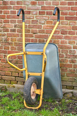 Garden wheelbarrow leaning against a red brick wall in a home backyard. Landscaping equipment and tools to carry and transport soil, manure, compost and fertilizer. Done cleaning a landscaped yardの写真素材