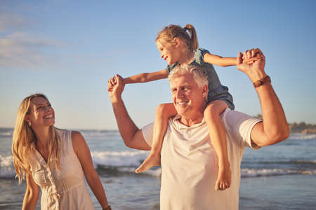 Closeup of a little caucasian girl being carried by her grandpa while her mother walks on the beach during sunset. Family fun in the summer sunの写真素材