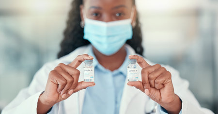 Medical doctor holding bottles of the covid cure. Ready to save the world from covid. African American doctor holding vials of the corona virus antidote.Healthcare professional wearing a maskの写真素材