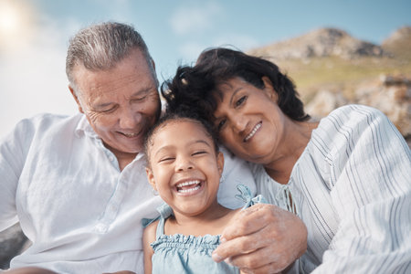Smiling mixed race grandparents sitting with granddaughter on a beach. Adorable, happy, hispanic girl bonding with grandmother and grandfather outside on weekend. Seniors and child enjoying free timeの写真素材