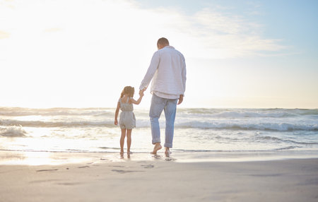 Rearview cute mixed race girl standing hand in hand with her father in the sea at the beach. A young man and his daughter holding hands while standing in the water and looking at the view at sunsetの写真素材