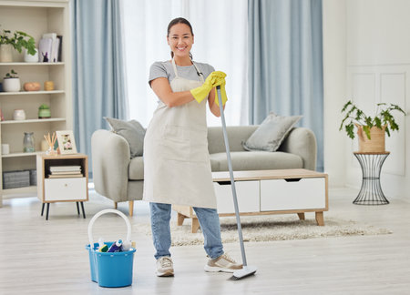 A happy smiling mixed race woman cleaning the floor of her apartment. One Asian woman using a broom and a bucket of cleaning supplies to clean the floorの写真素材