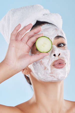 Beautiful young mixed race woman wearing a face mask peel and towel while holding cucumber slices....
