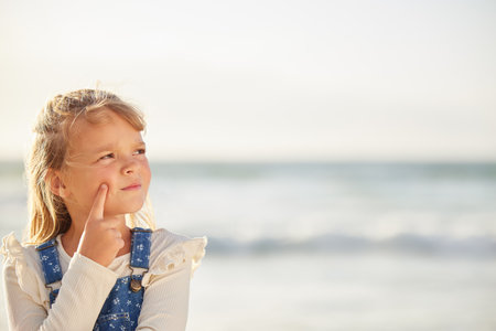 One adorable little girl thinking of ideas while standing on a beach outdoors. A cute young kid looking thoughtful while outside with the sea in the background. The mind and imagination on a childの写真素材