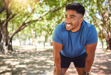 Happy athletic mixed race man smiling while standing with his hands on his knees in a park. Hispanic male resting after his run. Fit man pleased with his performance while exercising outdoorsの写真素材