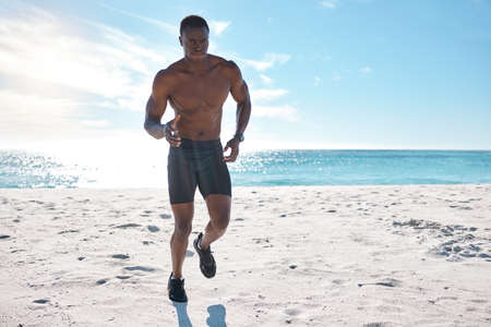 Fit young black man running and jogging on sand at the beach in the morning for exercise. One strong male bodybuilder athlete with six pack abs doing cardio workout to build muscle and enduranceの写真素材