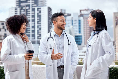 Two female doctors and a male colleague standing on the balcony outside a hospital smiling and having a conversation. mixed man and woman doctors talking to a caucasian female doctor while having a cup of coffee.の写真素材