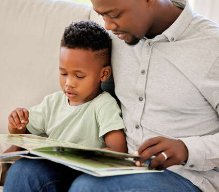 Young content African american father reading a book with his son sitting on the couch at home....