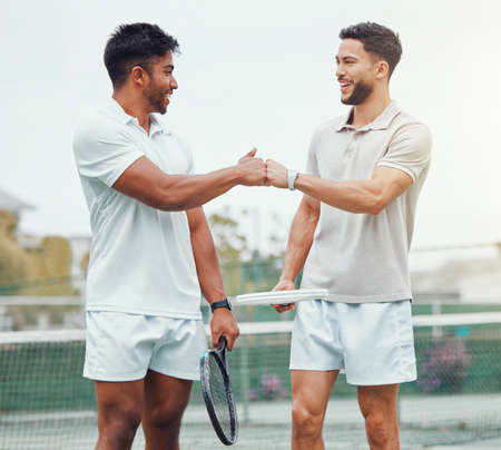 Two smiling ethnic tennis players giving fistbump with fist before playing court game. Fit athletes team standing and using hand gesture for good luck. Play competitive sports match for health fitnessの写真素材