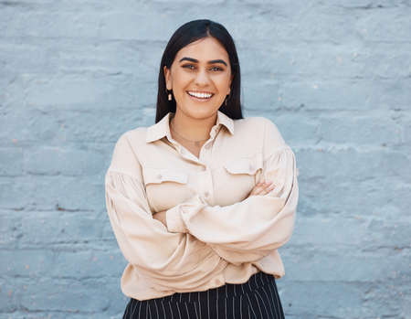 One young business woman of indian ethnicity standing outside against a grey wall with her arms crossed. Happy and confident mixed race executive looking positive and successful. Focus and ambitionの写真素材
