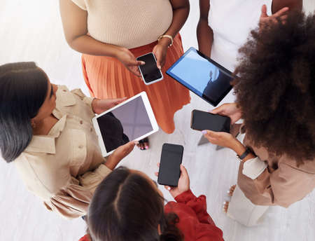 Above view of diverse group of unknown ambitious businesswomen huddled together and using technology in middle. Ethnic team of professional colleagues standing to   sync schedule in officeの写真素材