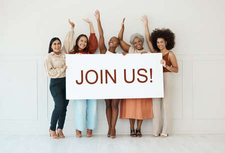 Group of five diverse young businesswomen standing against a wall in an office and holding a recruit sign. Happy colleagues holding a sign with a message while standing in a row together at workの写真素材