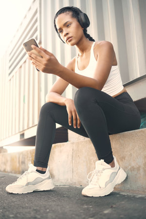 One fit young hispanic woman listening to music with headphones from a cellphone while taking a break from exercise outdoors. Female athlete texting and using fitness apps online while browsing social media and watching workout tutorials.の写真素材
