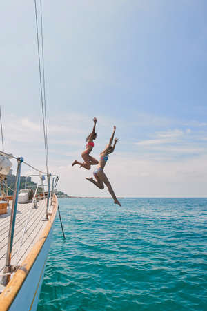 Two excited friends jumping off a boat into the ocean to swim during cruise. Two cheerful women in bikinis jumping off a boat to swim in the ocean together on holiday cruiseの写真素材