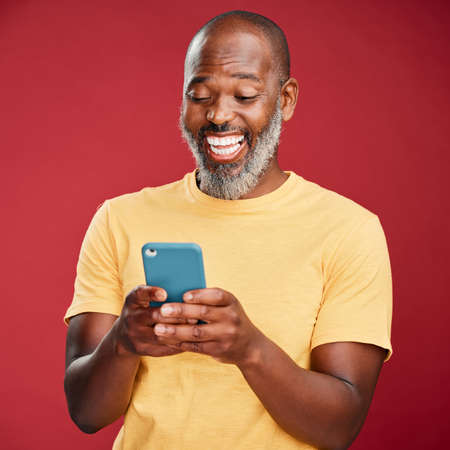 One happy African American man standing against a red studio background, holding and using his...