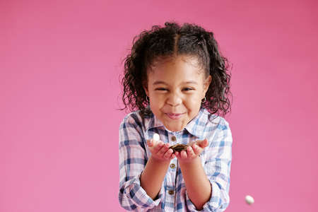 A pretty little mixed race girl with curly hair blowing confetti from her hands against a pink...